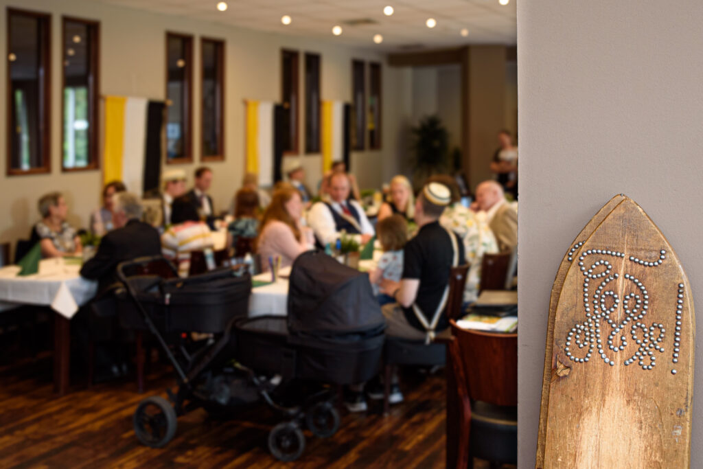 Im Vordergrund stehen zwei Schalgbretter mit dem Zirkel der Burschenschaft Vandalia. Im Hintergrund sieht man unschaf eine Gruppe von jungen und älteren Menschen sitzen.